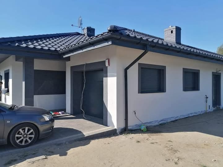 Nowoczesny dom jednorodzinny z białą elewacją i grafitowymi detalami, widoczny fragment samochodu i niezagospodarowany teren przed budynkiem.