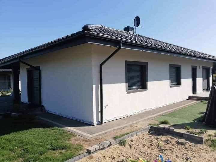 Nowoczesny dom jednorodzinny z jasną elewacją, ciemną dachówką i szarymi ramami okiennymi, widok z zewnątrz, z przygotowanym terenem wokół budynku i piaskownicą w rogu kadru.