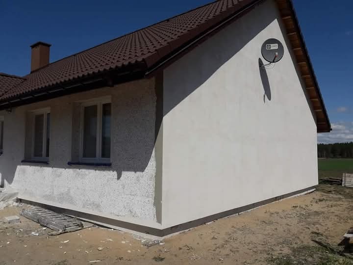 Fragment domu jednorodzinnego po nałożeniu nowej elewacji w kolorze białym, widoczna antena satelitarna, brązowy dach i fragment terenu przed budynkiem.