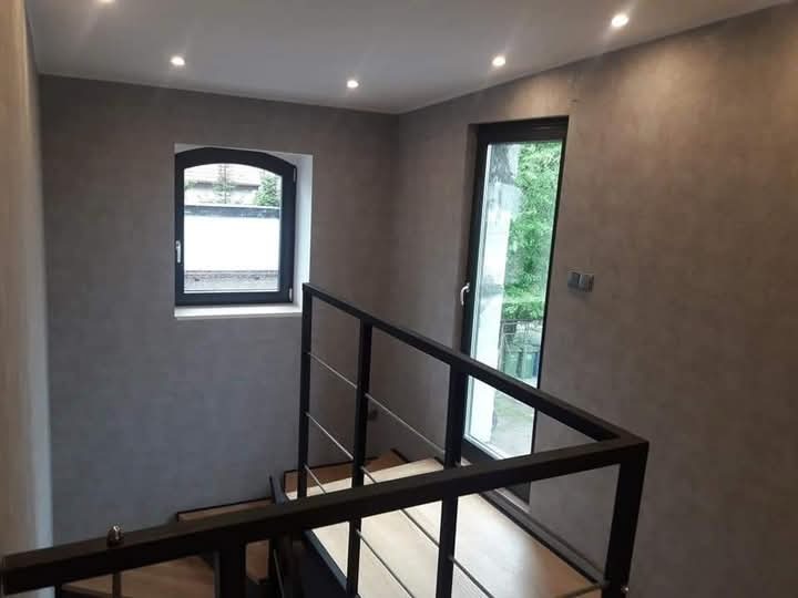 Klatka schodowa z drewnianymi schodami, czarną metalową balustradą i ścianami wykończonymi szarą, dekoracyjną farbą strukturalną. Widoczne dwa okna o różnych kształtach i oświetlenie punktowe...