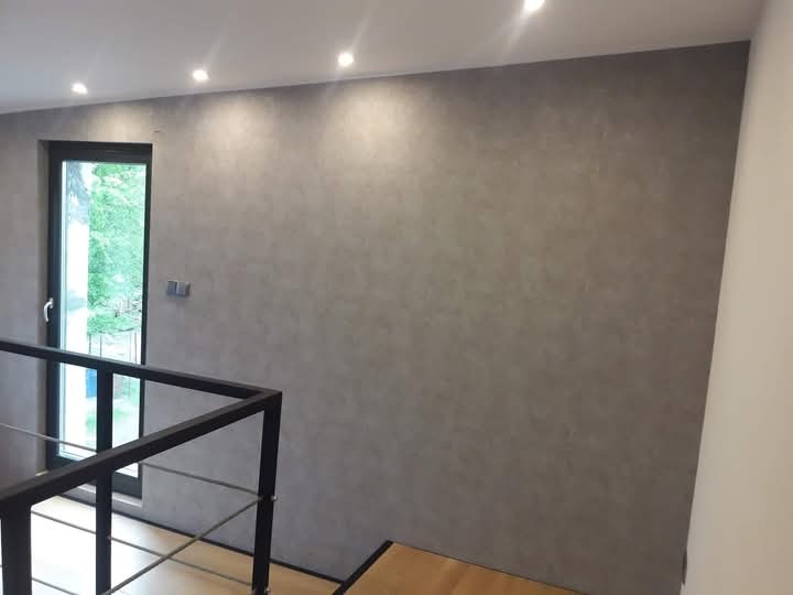 Ściana pokryta szarą, dekoracyjną powłoką, widok z góry schodów z czarną balustradą, okno z widokiem na zieleń na lewo od ściany, oświetlenie punktowe na suficie.