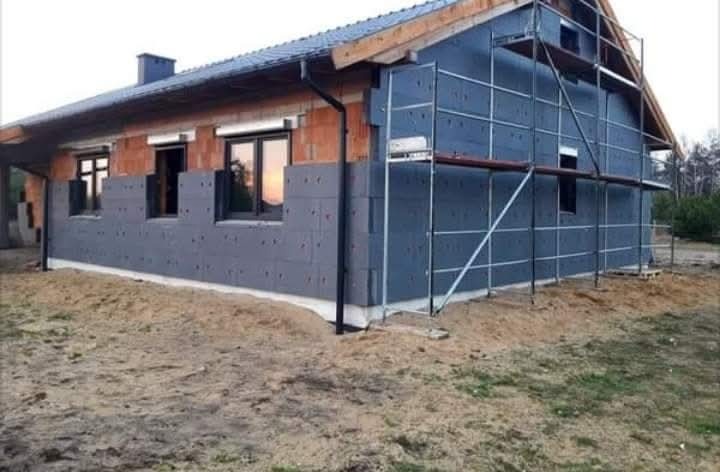 Dom w trakcie prac związanych z dociepleniem elewacji. Część ściany pokryta szarymi płytami izolacyjnymi, druga część w surowej cegle. Widoczne rusztowania.