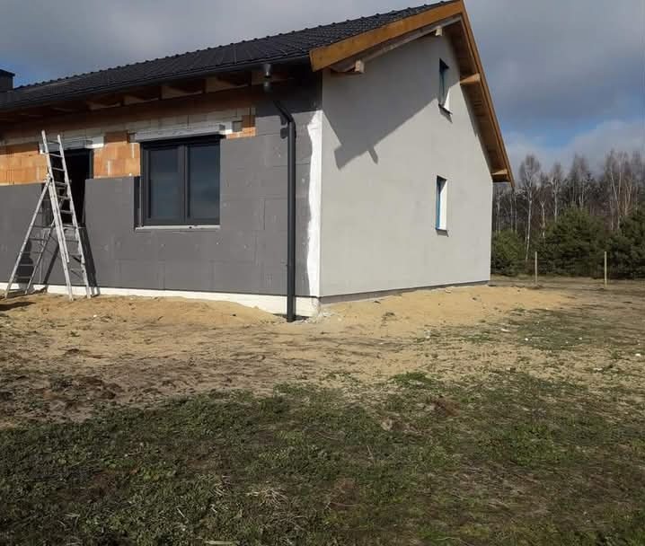 Dom jednorodzinny w trakcie ocieplania elewacji: fragment ściany z szarą izolacją styropianową, drabina oparta o ścianę, druga część ściany pomalowana na szaro, widoczne okno z czarną ramą...