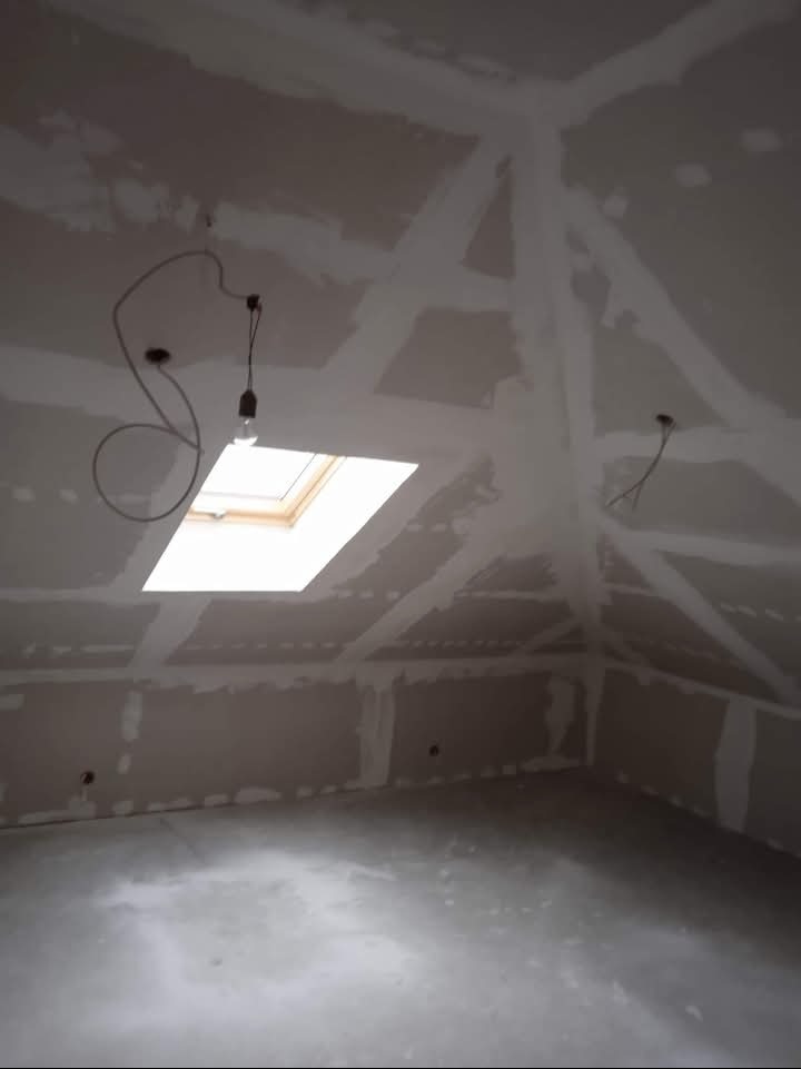 Poddasze w trakcie wykończenia płytami karton gips z widocznym oknem dachowym i prowizorycznym oświetleniem.