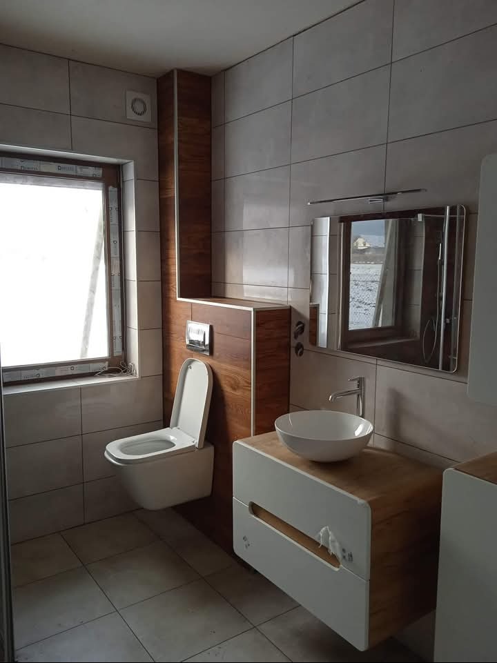 Nowoczesna łazienka z wiszącą toaletą, umywalką nablatową na szafce z drewnianymi akcentami i lustrem, wykończona płytkami w odcieniach szarości.