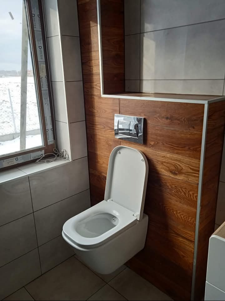 Wisząca toaleta z deską wolnoopadającą, zamontowana na ścianie wyłożonej płytkami imitującymi drewno, z chromowanym przyciskiem spłukującym. Widoczne płytki w odcieniach szarości i okno z widokiem...