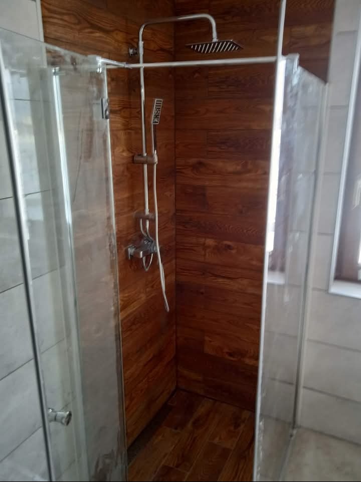 Kabina prysznicowa z drewnopodobnymi panelami ściennymi i deszczownicą, widoczna armatura i szklane drzwi, z delikatnym odblaskiem światła na powierzchni.