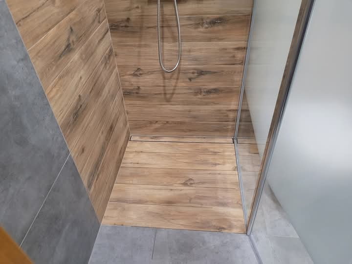 Kabina prysznicowa wyłożona płytkami imitującymi drewno, z odpływem liniowym i szklaną ścianką, kontrastująca z szarą posadzką, widok z góry.