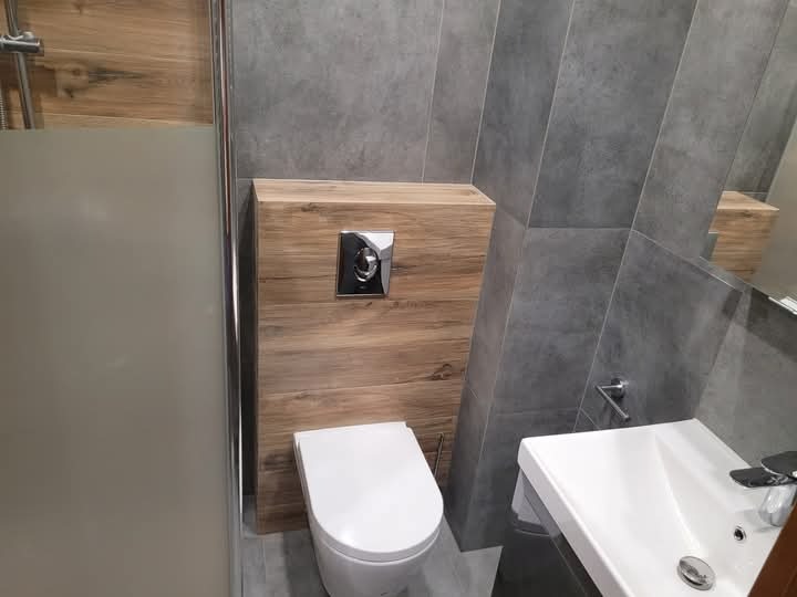 Nowoczesna łazienka z szarymi płytkami, drewnianą zabudową toalety, białą miską ustępową i umywalką, widoczna kabina prysznicowa z matową szybą.