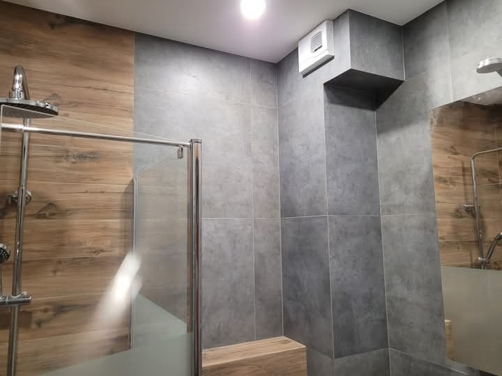 Wykończona łazienka z kabiną prysznicową, szare płytki na ścianach, imitacja drewna na fragmencie ściany, widoczny wentylator sufitowy.