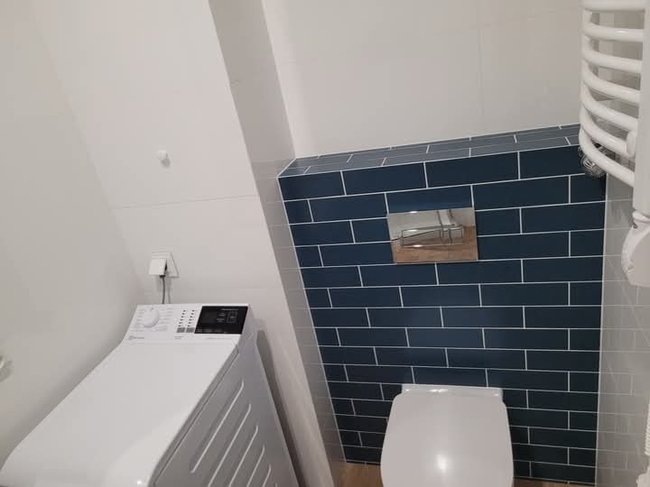 Ukończona łazienka na poddaszu ze skośnym sufitem, z ciemnymi płytkami ułożonymi w cegiełkę, podwieszaną toaletą, pralką i grzejnikiem drabinkowym.