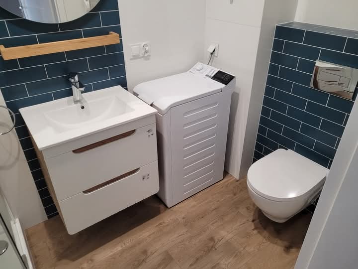 Nowoczesna łazienka z umywalką na szafce z drewnianymi akcentami, pralką ładowaną od góry i podwieszaną toaletą na tle ściany wyłożonej ciemnymi, prostokątnymi płytkami.