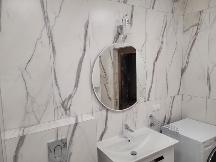 Nowoczesna łazienka z białymi płytkami imitującymi marmur, okrągłe lustro z oświetleniem, biała umywalka z baterią oraz pralka.