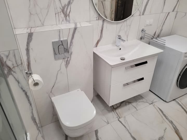 Nowoczesna łazienka wykończona białymi płytkami imitującymi marmur, z wiszącą toaletą, szafką z umywalką i pralką, całość utrzymana w jasnej kolorystyce.