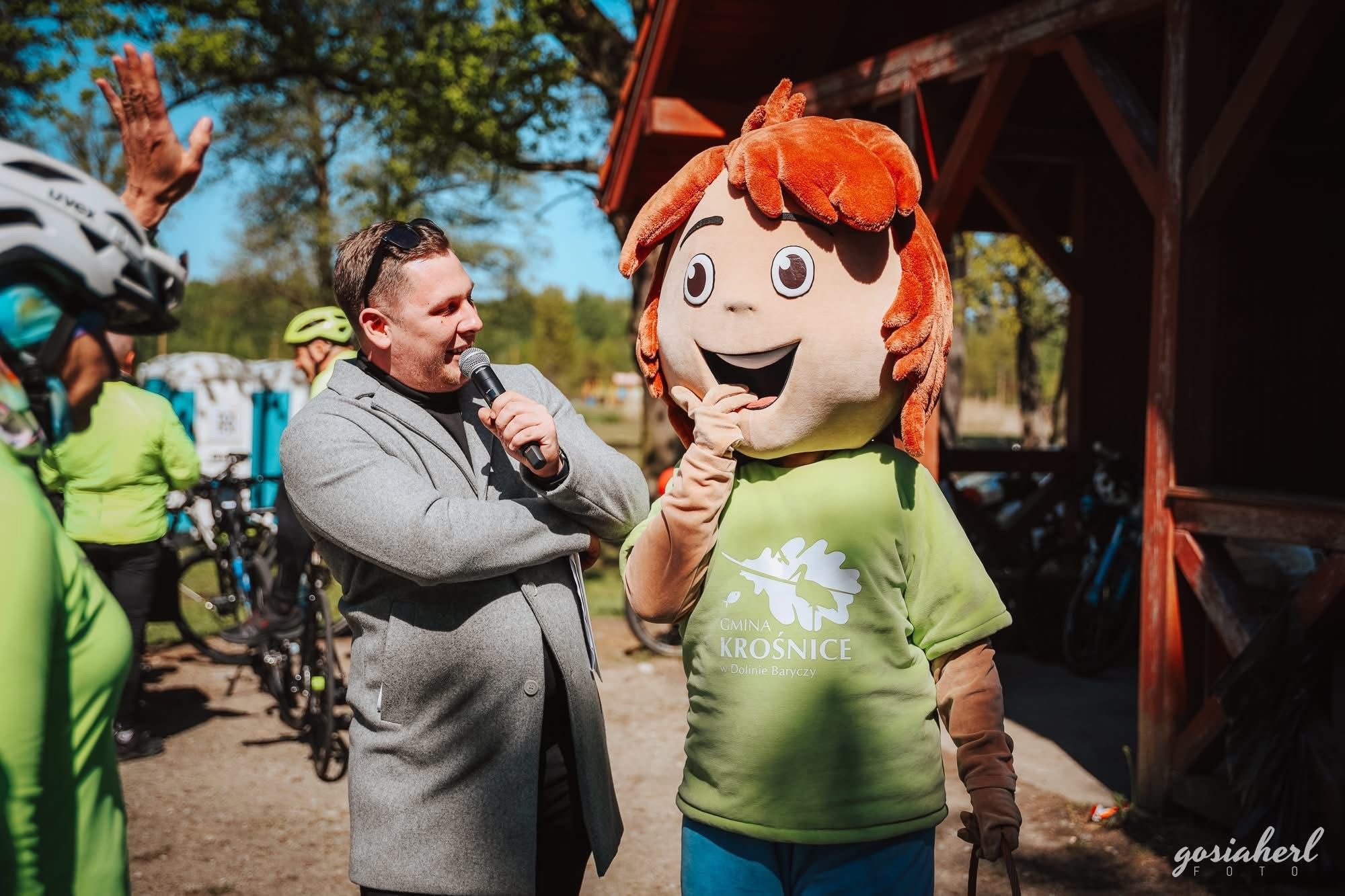 Mężczyzna w szarym płaszczu przemawia do maskotki Gminy Krośnice podczas wydarzenia plenerowego. W tle rowery i uczestnicy.
