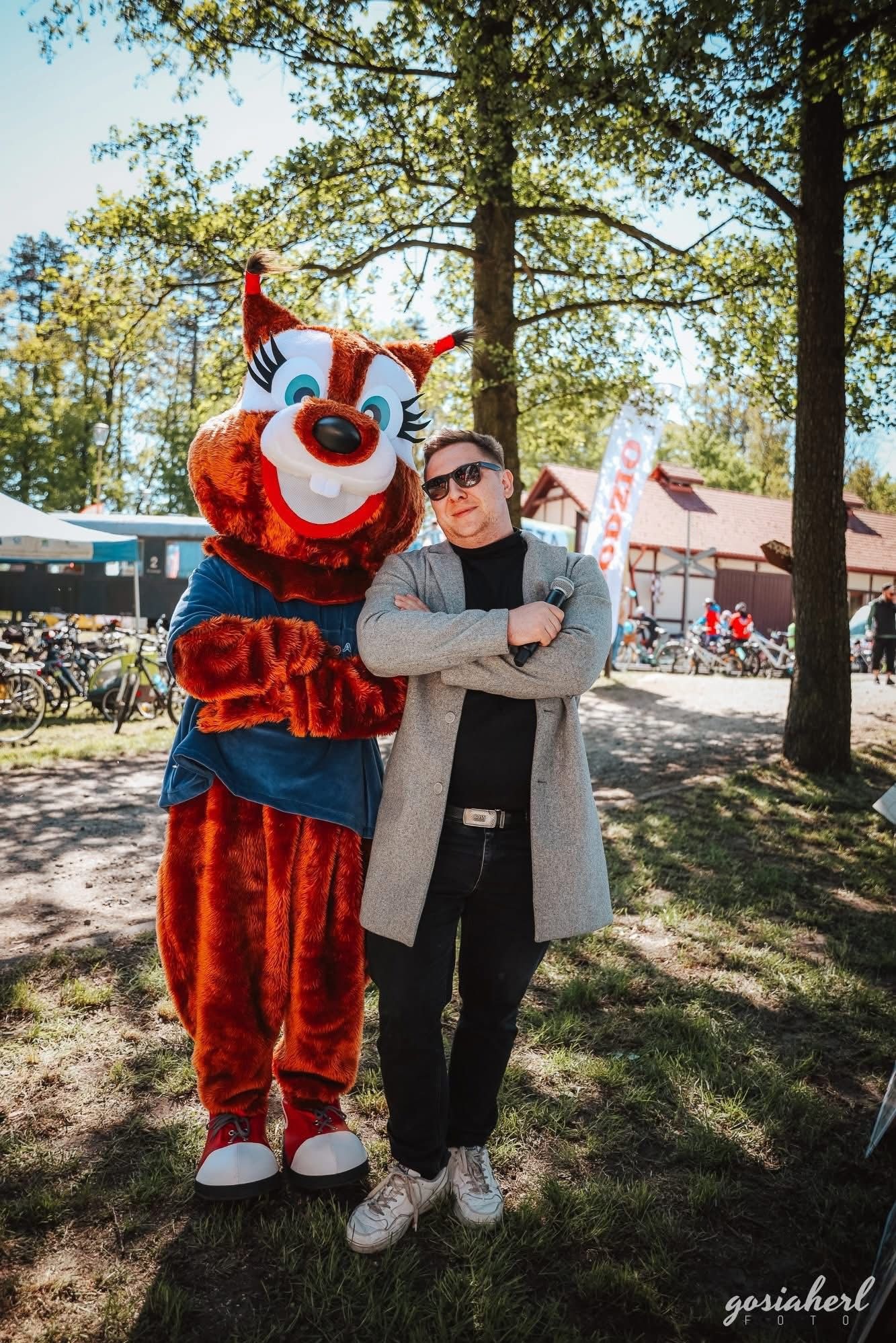 Mężczyzna w okularach, z mikrofonem, pozuje z animowaną maskotką w plenerze, prawdopodobnie podczas imprezy plenerowej. W tle rowery i budynek.