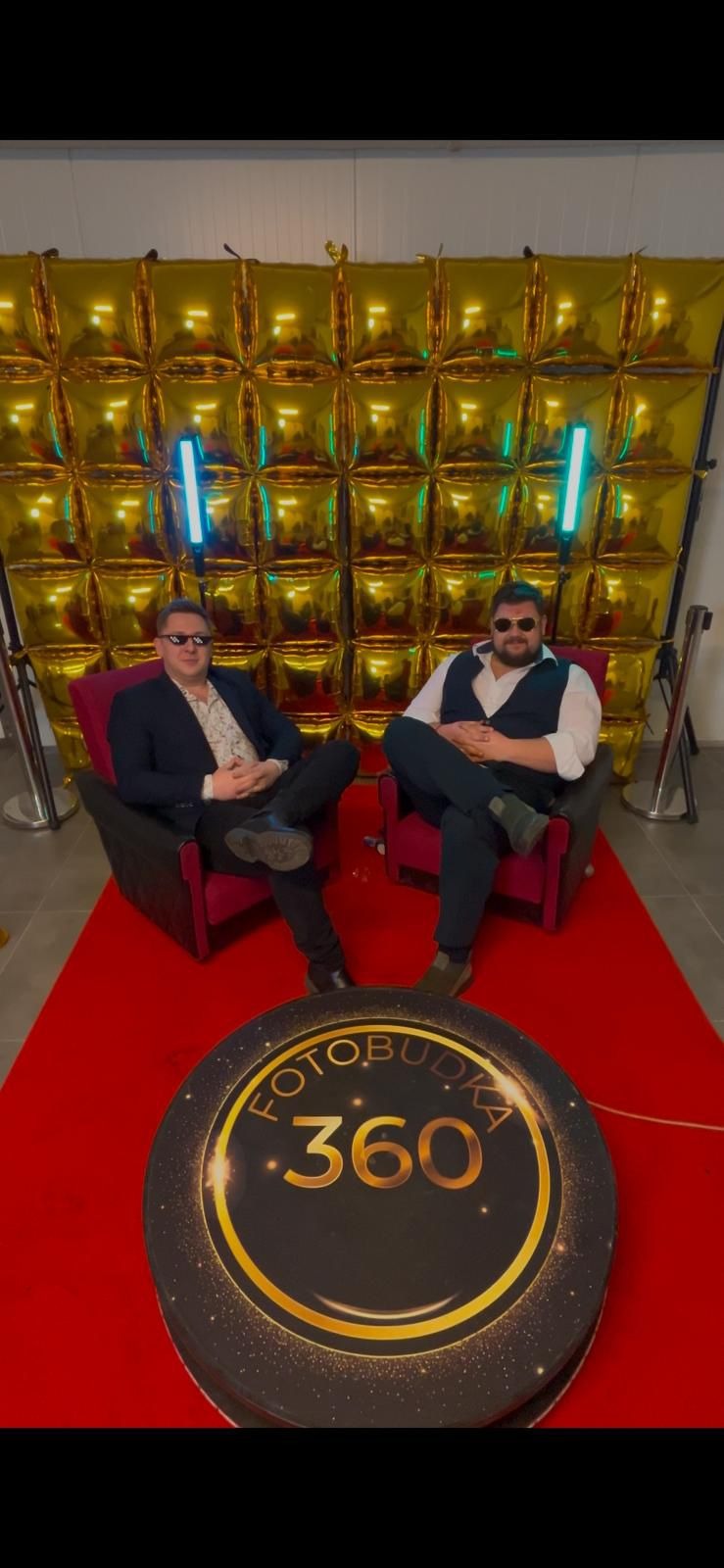 Dwóch mężczyzn w fotobudce 360 na czerwonym dywanie przed złotą ścianką z balonów i neonowym oświetleniem. Panowie w eleganckich strojach pozują w fotobudce.