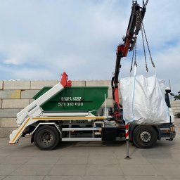 GRUPA KOSZ SPÓŁKA Z OGRANICZONĄ ODPOWIEDZIALNOŚCIĄ - Ciężarówka z hakiem i zielonym kontenerem podnosi duży biały worek z gruzem na tle betonowych bloków.