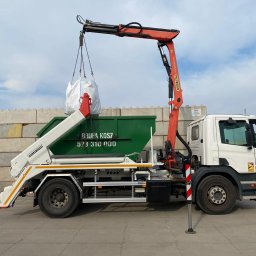 GRUPA KOSZ SPÓŁKA Z OGRANICZONĄ ODPOWIEDZIALNOŚCIĄ - Ciężarówka z pomarańczowym dźwigiem Palfinger podnosi biały worek nad zielonym kontenerem. Widoczny numer telefonu na kontenerze i betonowy mur w tle.