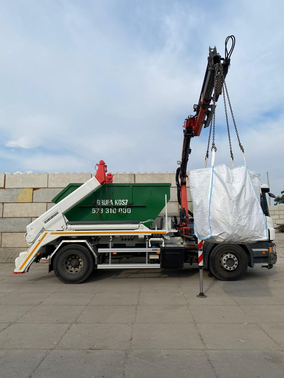 Ciężarówka z hakiem i zielonym kontenerem podnosi duży biały worek z gruzem na tle betonowych bloków.