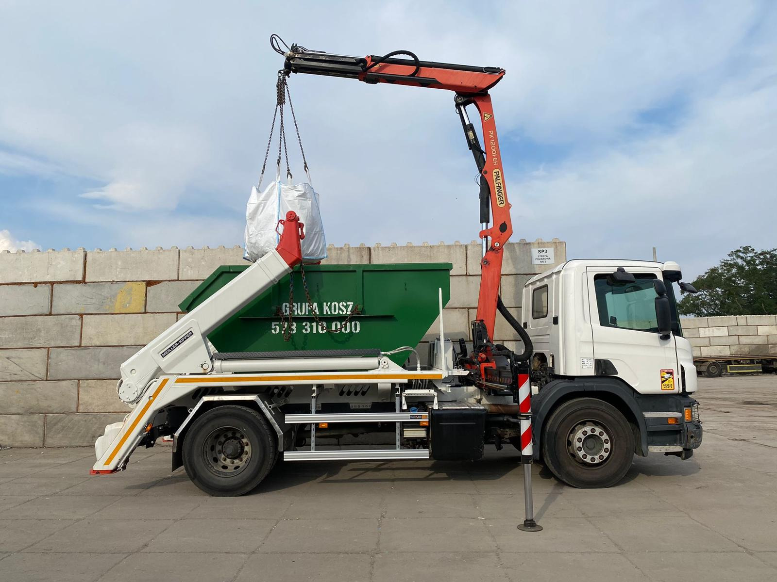 Ciężarówka z pomarańczowym dźwigiem Palfinger podnosi biały worek nad zielonym kontenerem. Widoczny numer telefonu na kontenerze i betonowy mur w tle.