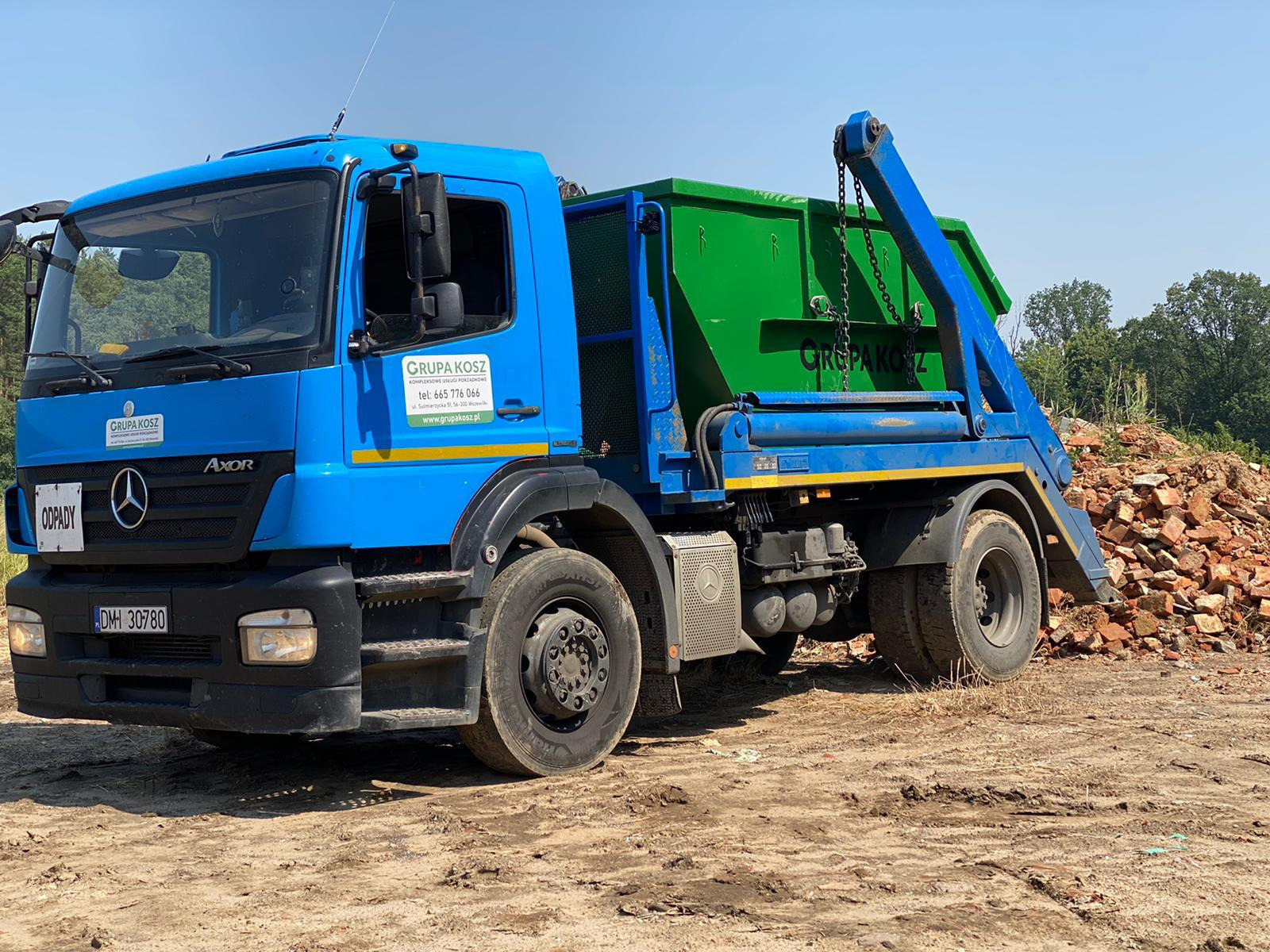 Niebieska ciężarówka z zielonym kontenerem na podwoziu, zaparkowana na ziemi obok sterty gruzu ceglanego, z logo firmy 'Grupa Kosz' widocznym na kabinie.