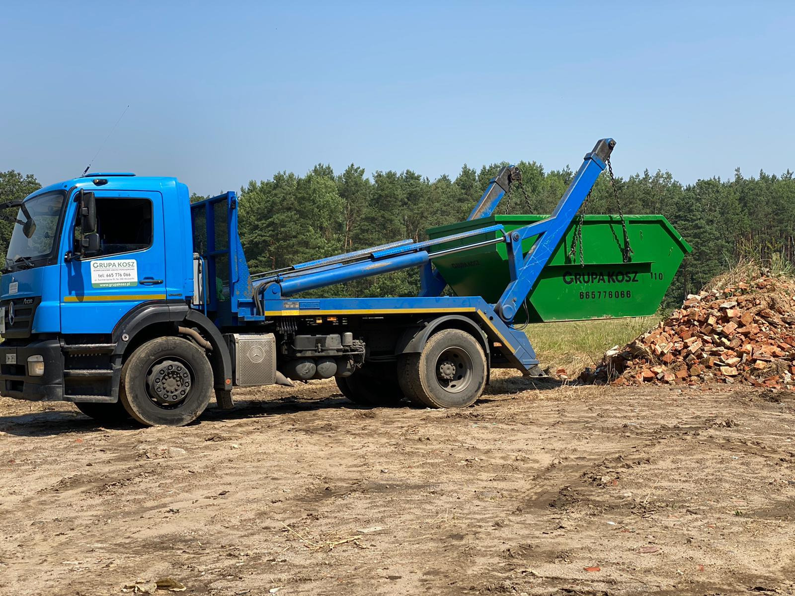 Niebieska ciężarówka z zielonym kontenerem, firmy ze Stawca, w trakcie załadunku gruzu ceglanego. Widoczne logo firmy na pojeździe.