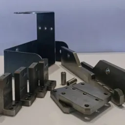 Zestaw precyzyjnie wykonanych, metalowych elementów o różnych kształtach i rozmiarach, w tym kątowniki z otworami, płyty z rowkiem i tuleje, ułożonych na białym tle.