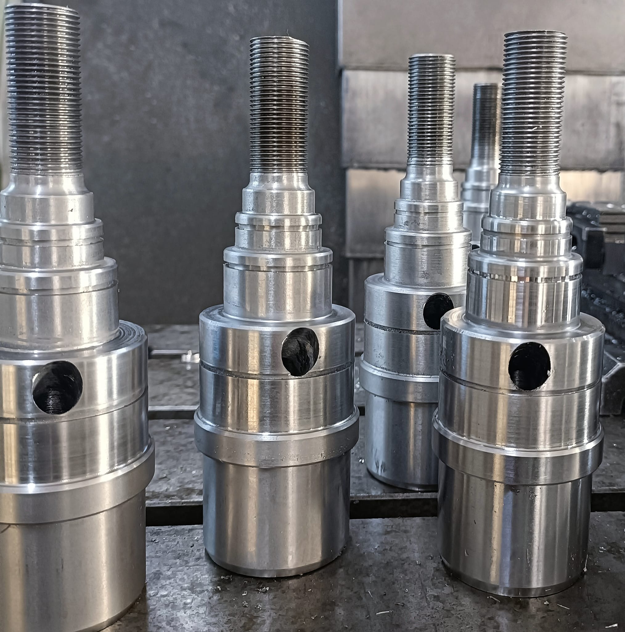 Szereg gotowych, metalowych elementów z gwintem i otworem, po obróbce, leżących na stole warsztatowym.