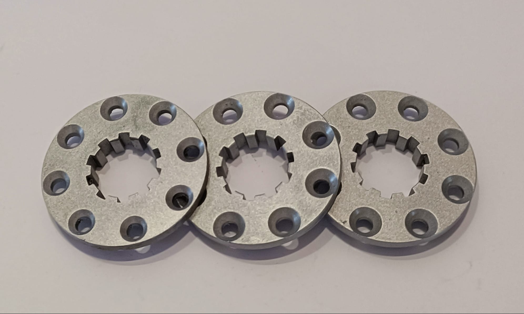 Trzy identyczne, metalowe pierścienie z ząbkowanym otworem centralnym i sześcioma otworami obwodowymi, ułożone blisko siebie na gładkim, jasnym tle, ukazujące precyzję obróbki CNC.