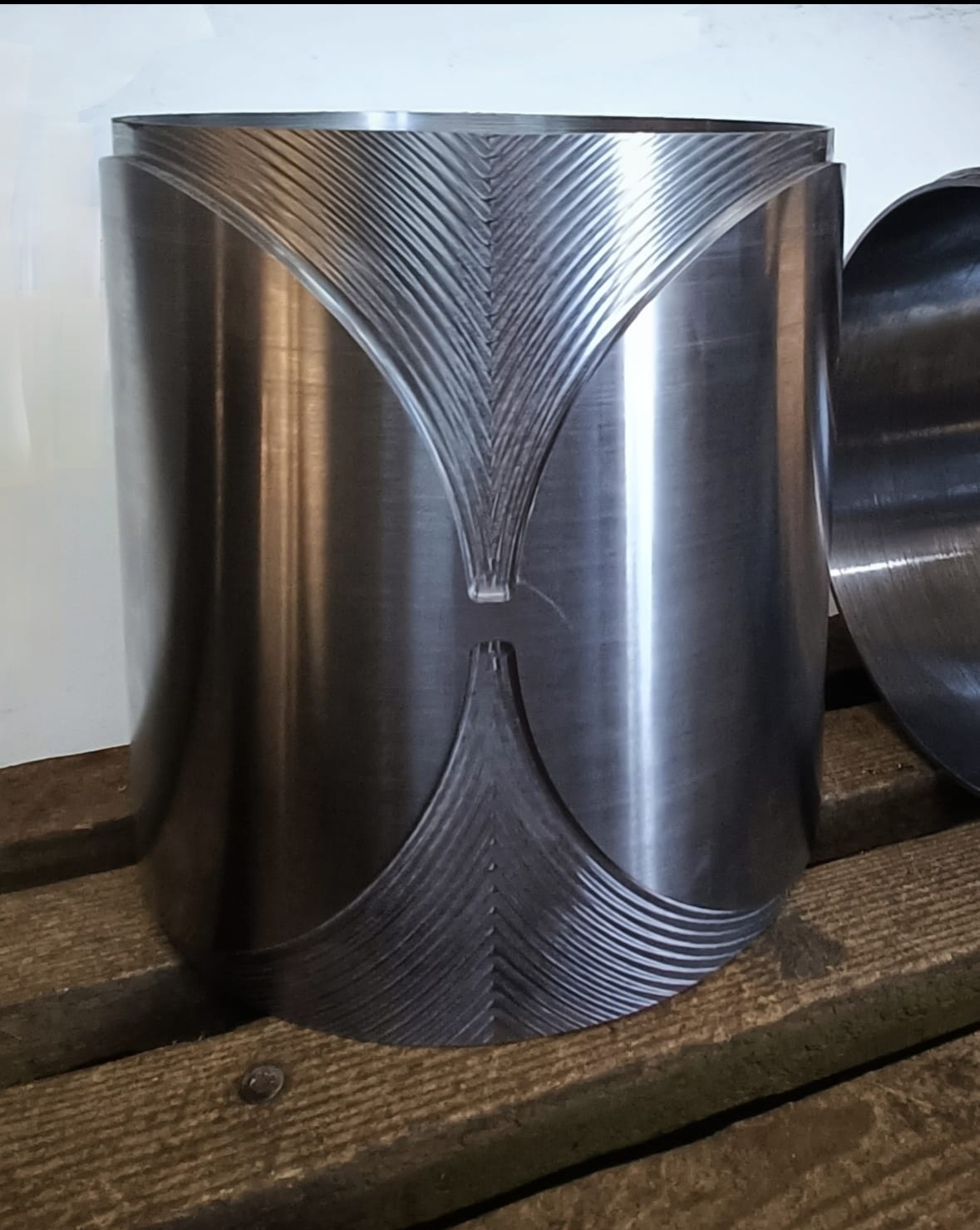 Metalowy cylinder z precyzyjnie wyfrezowanymi, symetrycznymi rowkami w kształcie klepsydry, leżący na drewnianej palecie.