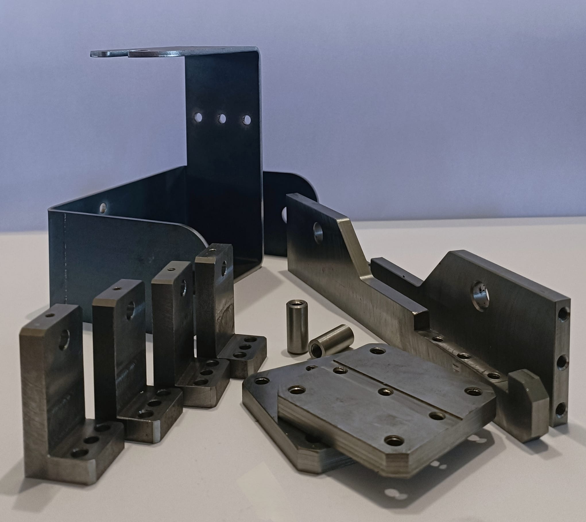 Zestaw precyzyjnie wykonanych, metalowych elementów o różnych kształtach i rozmiarach, w tym kątowniki z otworami, płyty z rowkiem i tuleje, ułożonych na białym tle.