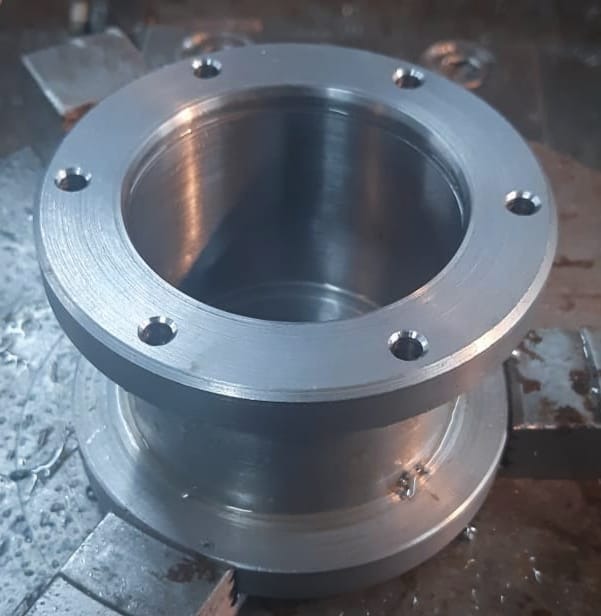 Szary metalowy element z kołnierzem i otworami na śruby, prawdopodobnie wytoczony, leżący na warsztatowym stole z widocznymi wiórami.