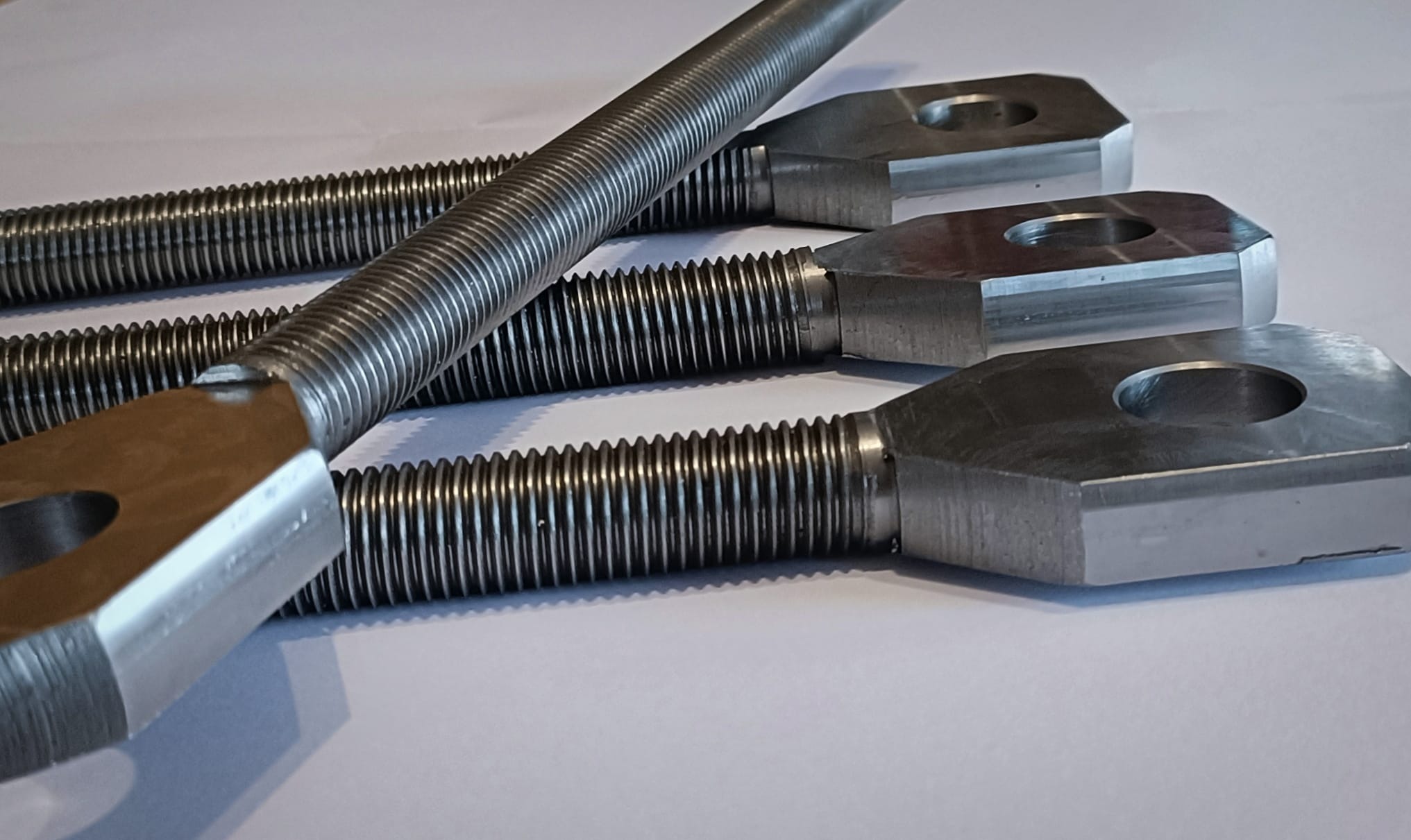 Trzy metalowe śruby z gwintem trapezowym i oczkami, ułożone równolegle na białym tle, widoczne ślady obróbki.