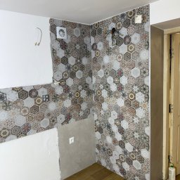 Janusz Brawiak Usługi Budowlane - Remont łazienki w trakcie: częściowo ułożone heksagonalne płytki na ścianie, widoczne kable, wentylator i fragment podłogi z paneli. Prace wykończeniowe w toku.