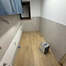 Janusz Brawiak Usługi Budowlane - Wnętrze łazienki w trakcie prac: położone płytki imitujące drewno na podłodze, częściowo wykończone ściany z szarych płytek, widoczne instalacje hydrauliczne i elektryczne.