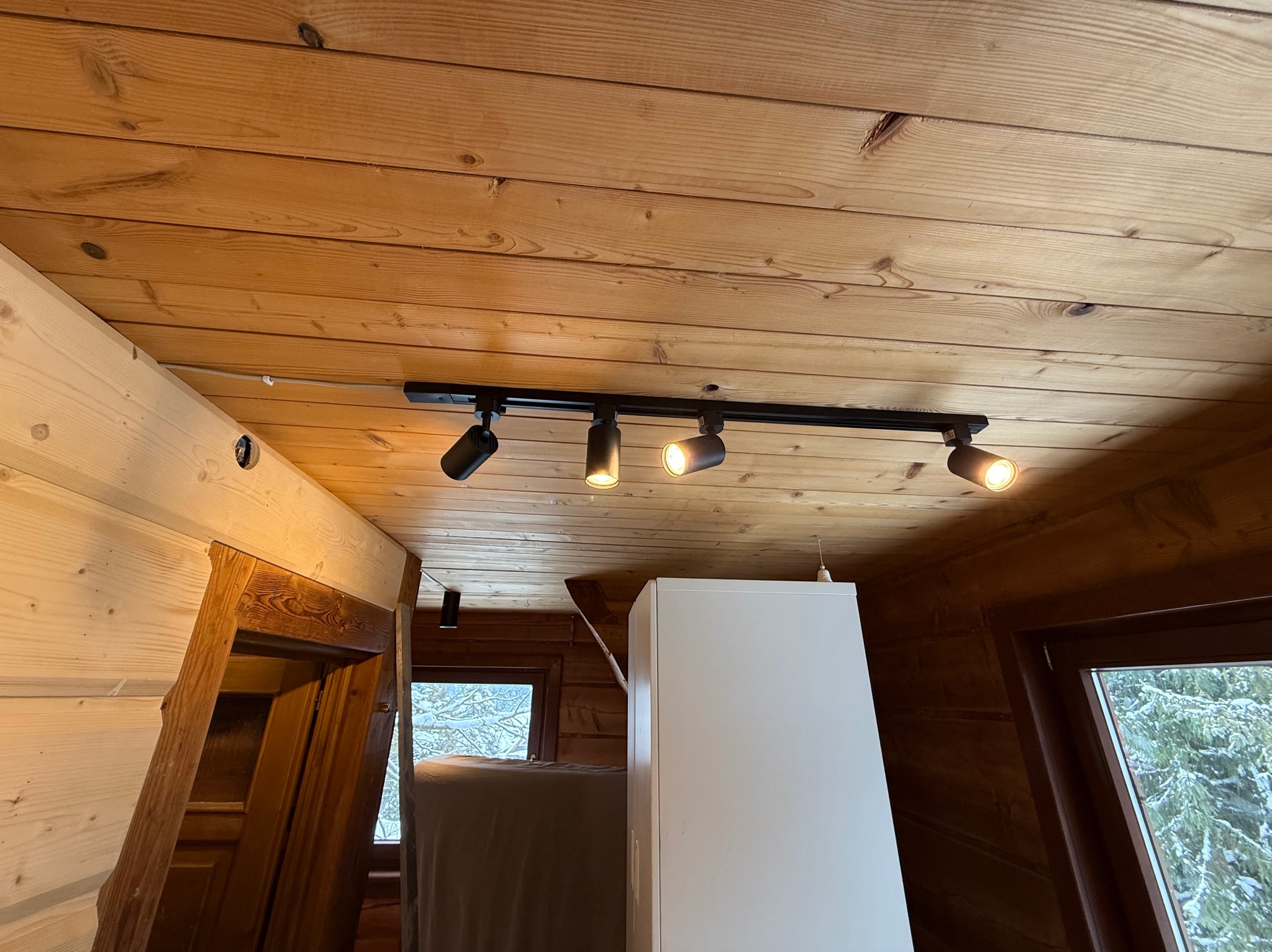 Drewniany sufit z zamontowanym czarnym systemem oświetleniowym z czterema reflektorami. Widoczne okno z widokiem na zaśnieżone drzewa i fragment białej szafki.