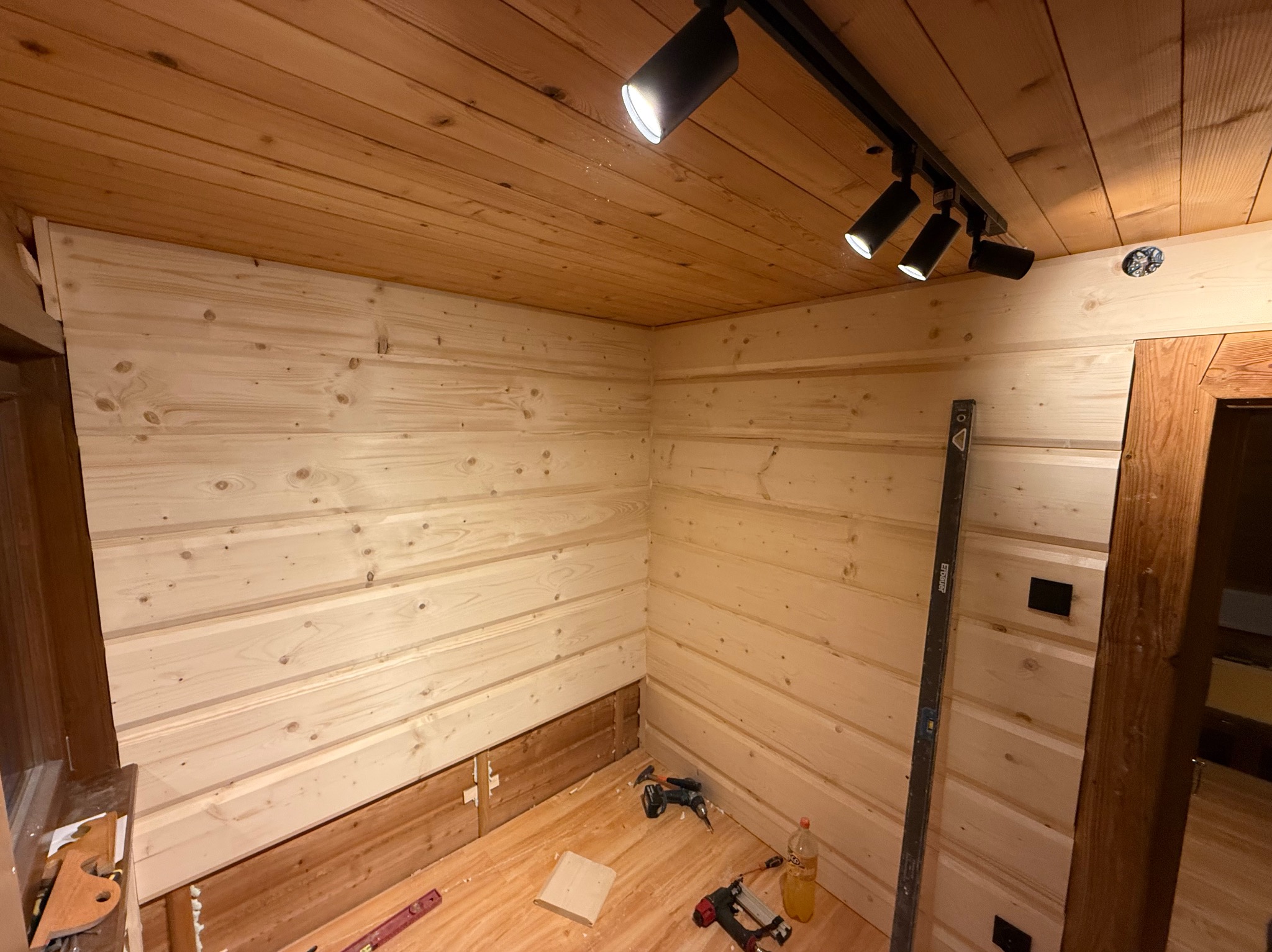 Wnętrze pokoju w trakcie wykańczania drewnem: boazeria na ścianach, surowa podłoga, narzędzia pracy i oświetlenie punktowe na suficie. Widoczny poziomica.