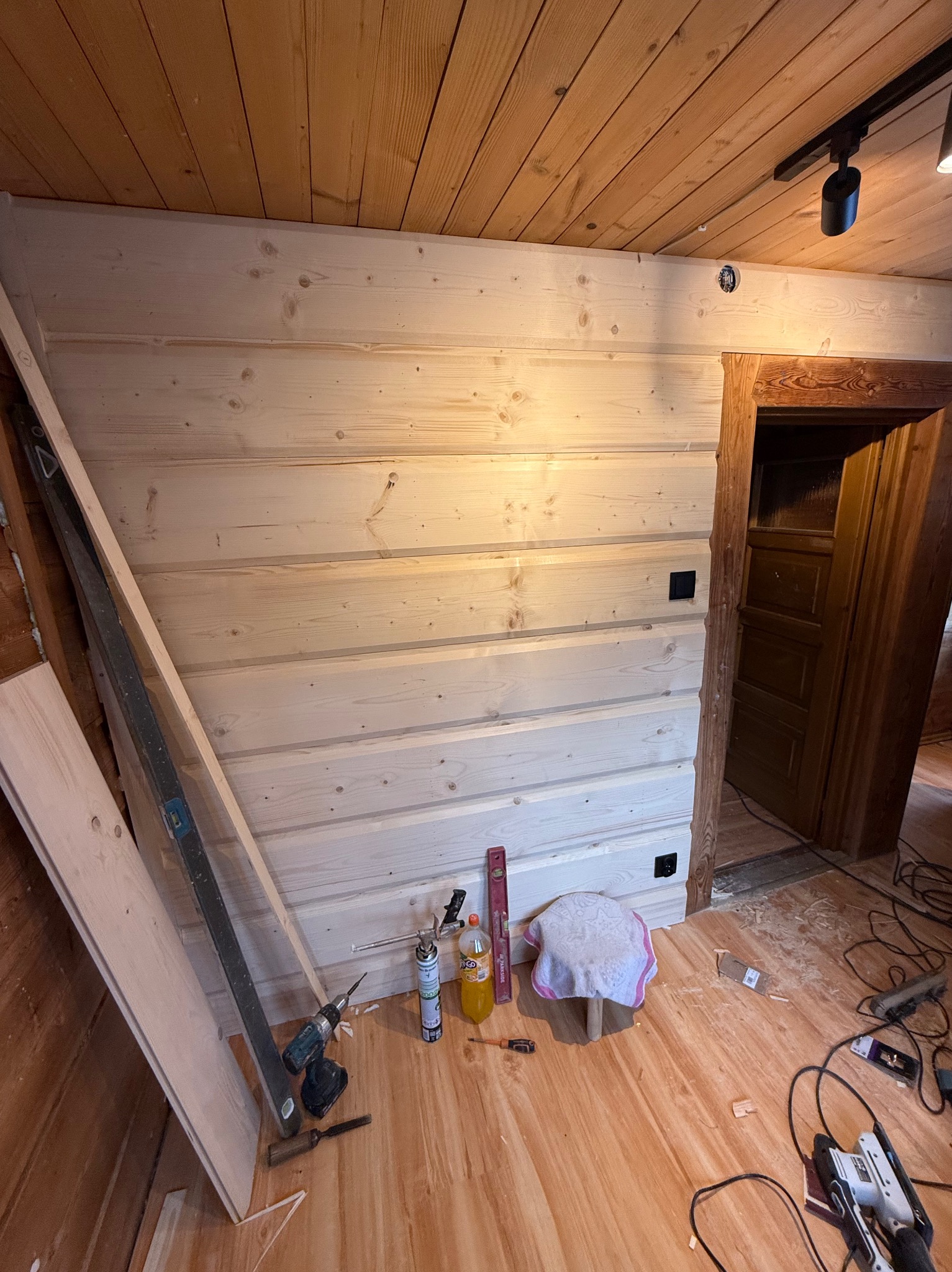 Ściana i sufit wykończone jasnym drewnem boazeryjnym, widoczne narzędzia pracy, poziomica, wiertarka, pianka montażowa. Fragment drewnianej futryny drzwiowej.