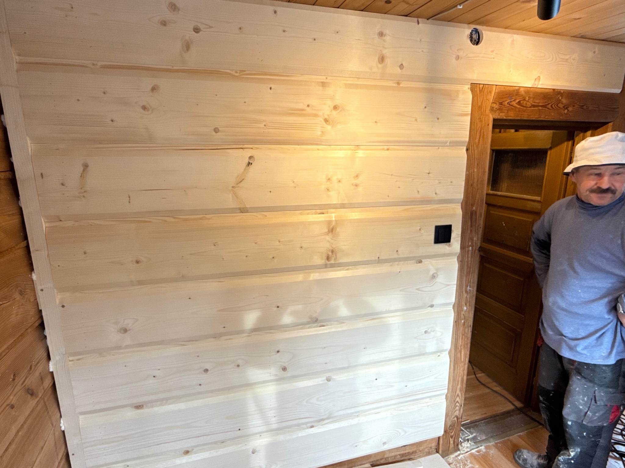 Ściana wyłożona jasnymi, drewnianymi panelami boazeryjnymi, z widocznym fragmentem drewnianej futryny drzwi i majsterkowiczem w tle. Widoczne łączenia paneli.