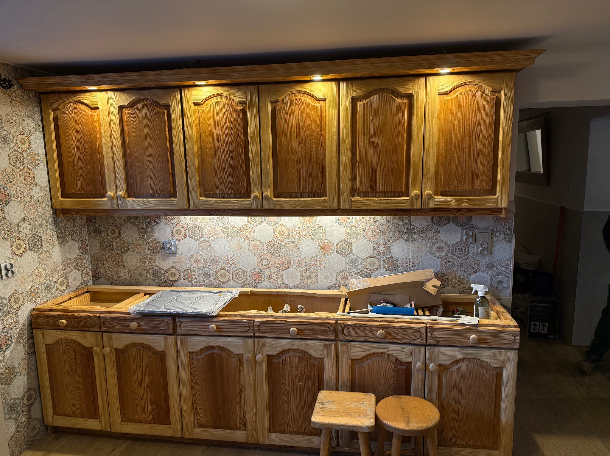 Drewniane szafki kuchenne z oświetleniem podszafkowym i wzorzystą mozaiką na ścianie, widoczne akcesoria montażowe i dwa stołki barowe.