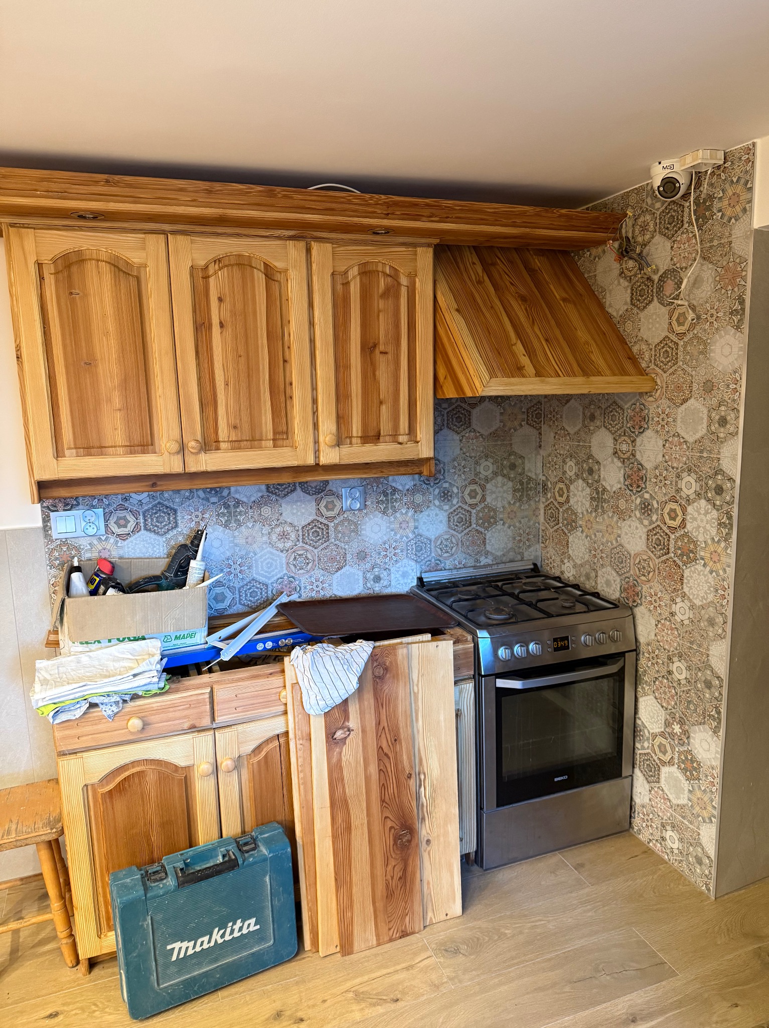 Kuchnia w trakcie remontu: drewniane szafki, heksagonalne płytki, kuchenka. Widoczne narzędzia i materiały budowlane. Ujęcie z bliska, ukazujące etap prac.