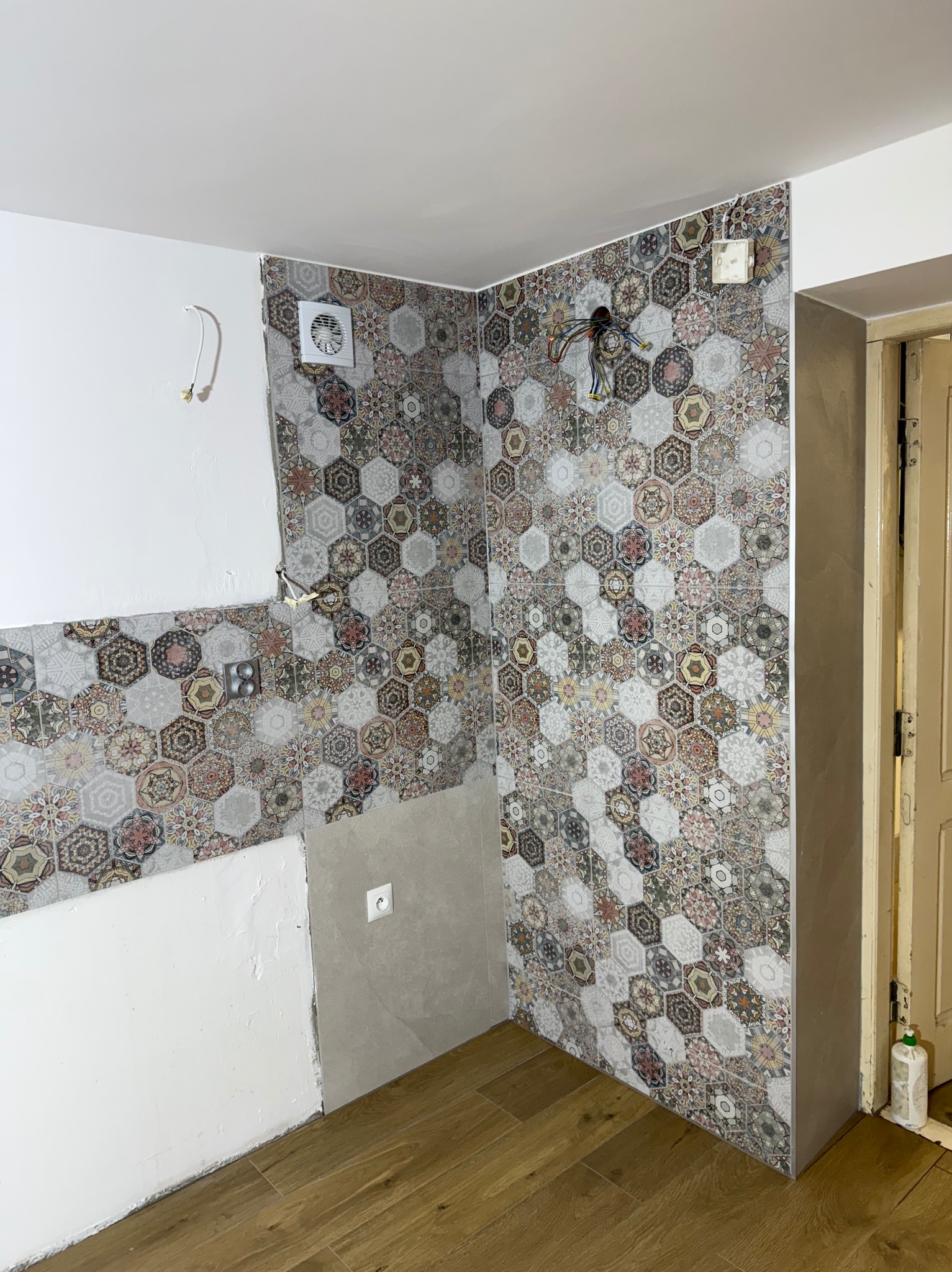 Remont łazienki w trakcie: częściowo ułożone heksagonalne płytki na ścianie, widoczne kable, wentylator i fragment podłogi z paneli. Prace wykończeniowe w toku.
