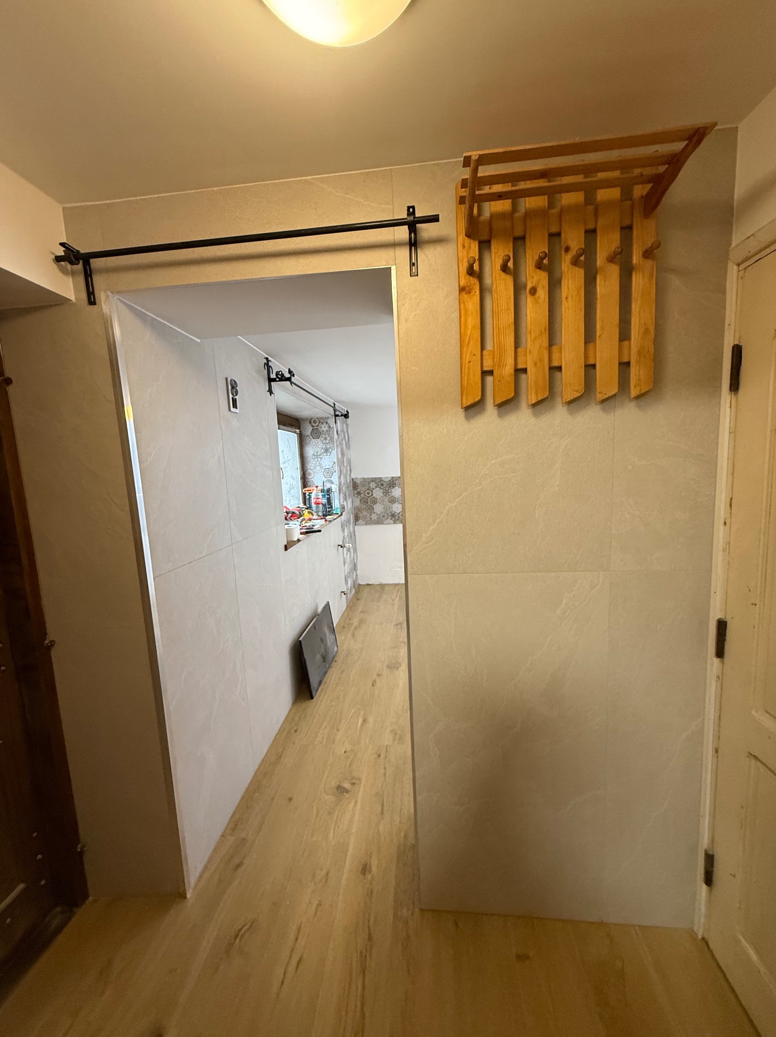 Wykończony korytarz z drewnianą podłogą i jasnymi płytkami na ścianach. Widoczny wieszak na ubrania i przesuwne drzwi do kuchni z oknem.