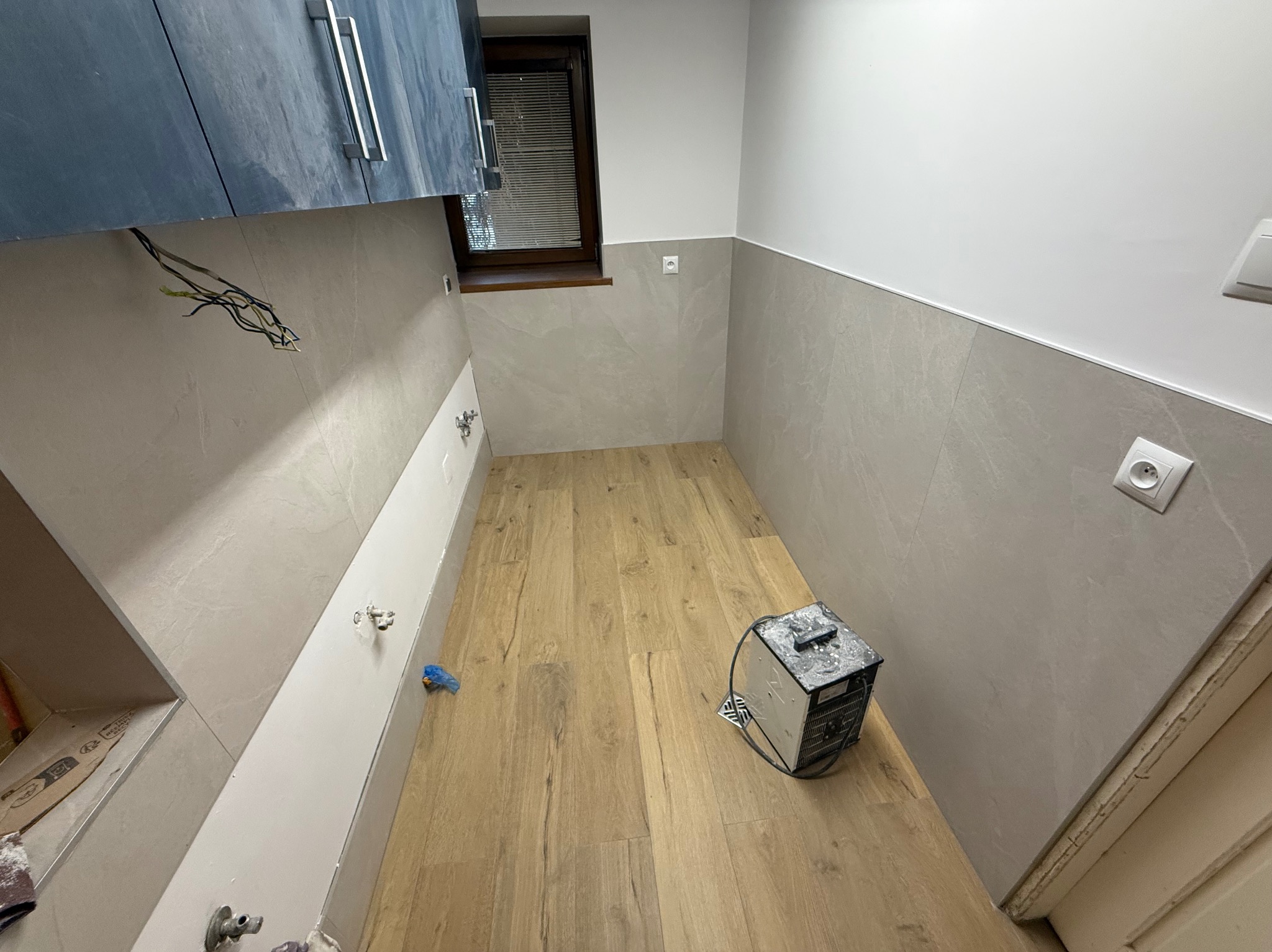 Wnętrze łazienki w trakcie prac: położone płytki imitujące drewno na podłodze, częściowo wykończone ściany z szarych płytek, widoczne instalacje hydrauliczne i elektryczne.