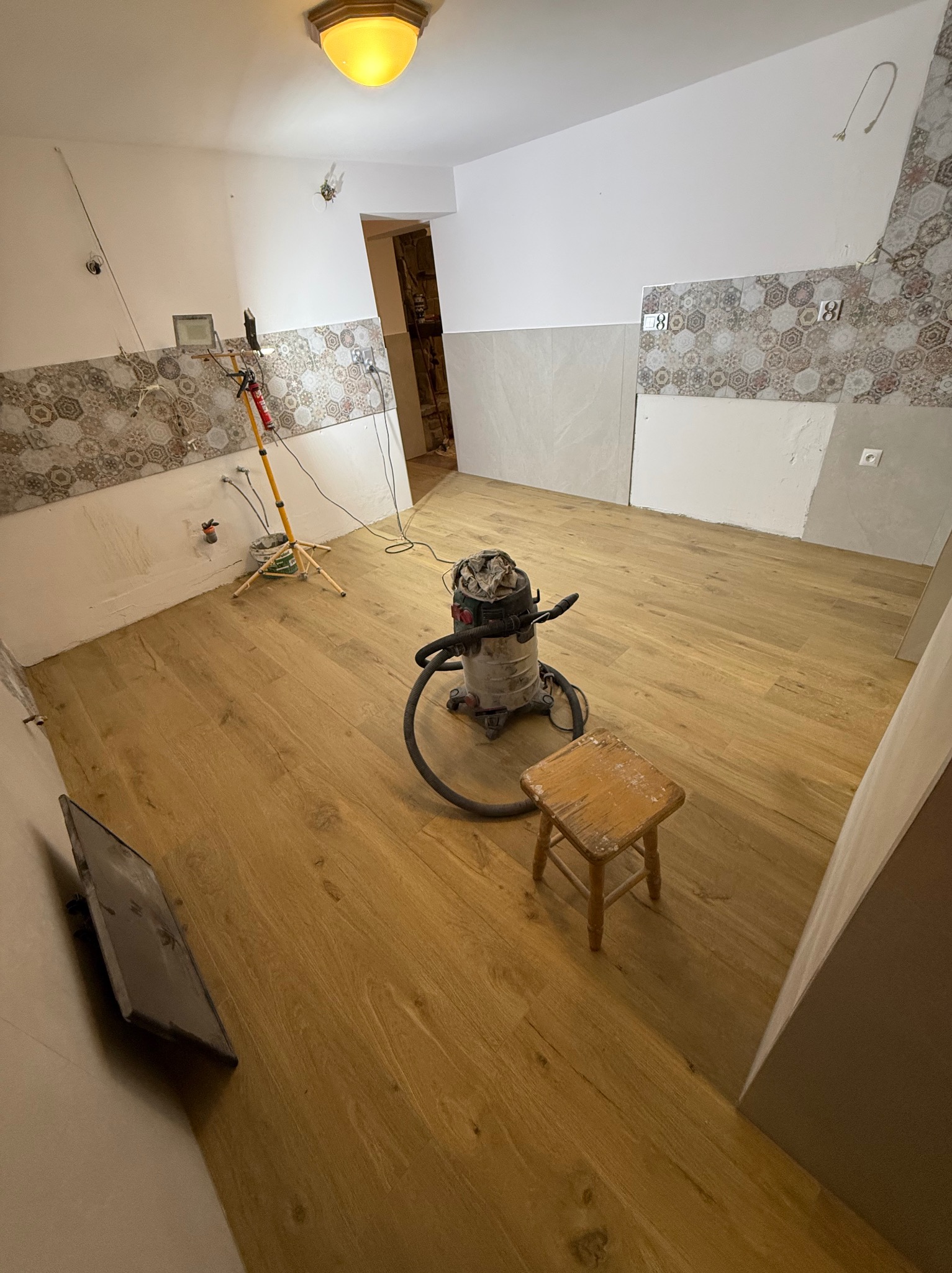 Remont pokoju: drewniana podłoga, częściowo położone płytki na ścianach, odkurzacz przemysłowy i stołek. Widoczne niedokończone prace i narzędzia.
