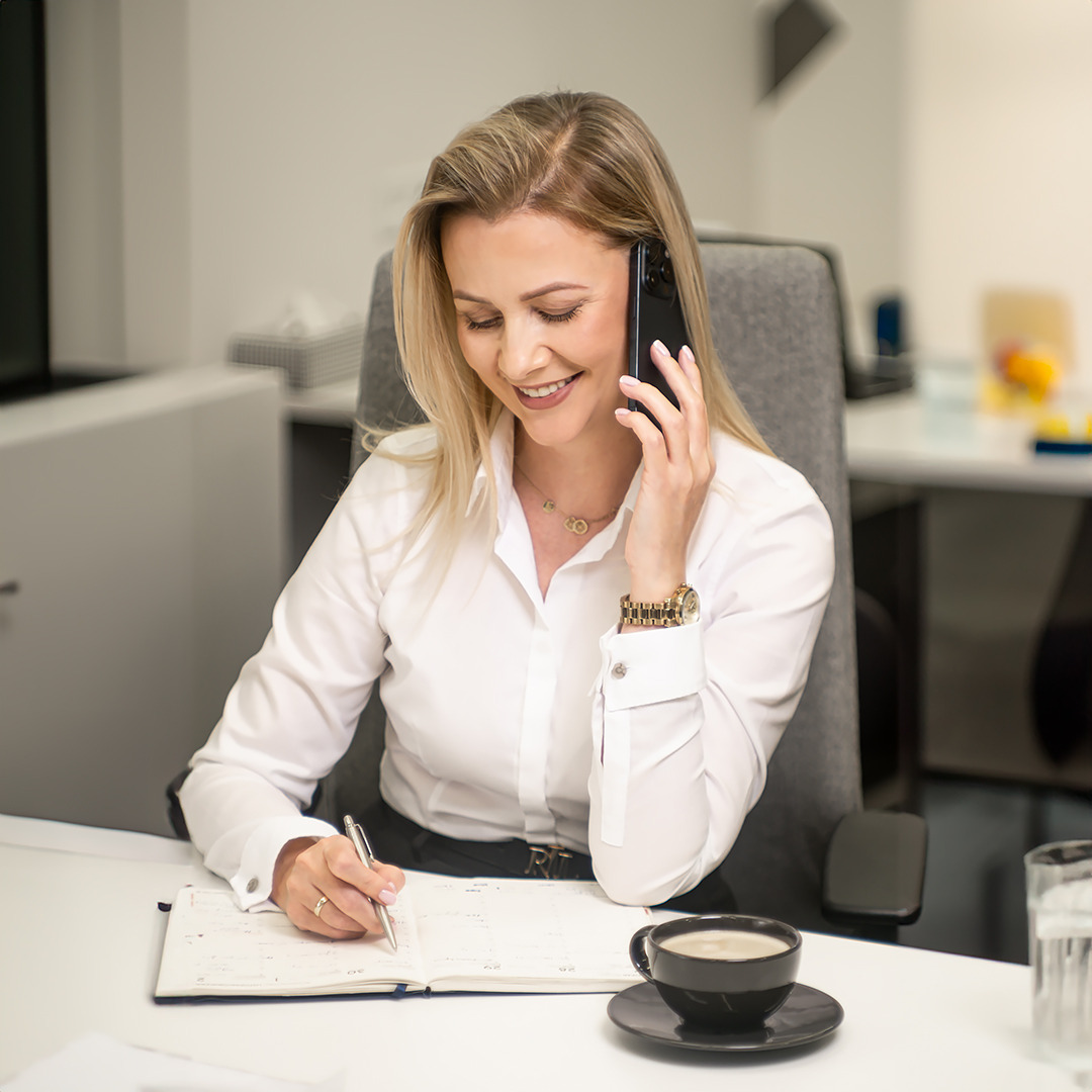 Blondwłosa kobieta w białej koszuli siedzi przy biurku, rozmawia przez telefon i notuje coś w kalendarzu. Na biurku stoi filiżanka kawy i szklanka wody.