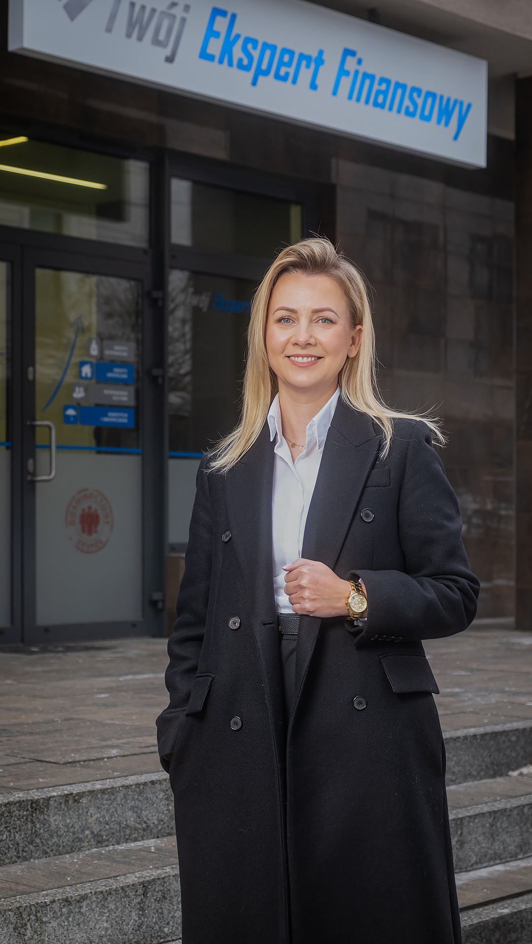 Profesjonalna blondynka w czarnym płaszczu, stojąca przed budynkiem z napisem 'Twój Ekspert Finansowy' w tle, uśmiechnięta, z zegarkiem na ręce.