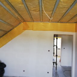 Prace Wykończeniowe - Poddasze w trakcie ocieplania, widoczne metalowe profile, wełna mineralna, żółta folia paroizolacyjna przy ścianach, oraz fragment ściany z przygotowanymi otworami pod gniazdka elektryczne.