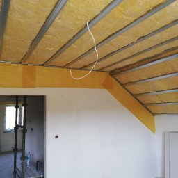 Prace Wykończeniowe - Poddasze w trakcie ocieplania, widoczne profile metalowe, żółta wełna mineralna, folia paroizolacyjna, przewód elektryczny zwisający z sufitu, fragment rusztowania w tle.